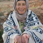 A woman in peasant garb with a woollen Berber jacket smiles and points to her blue fingertips.
