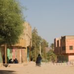 The silhouette of an Aït Atta woman wrapped in a black veil embroidered with bright wool in an earthen street lined with mud houses and acacia trees.