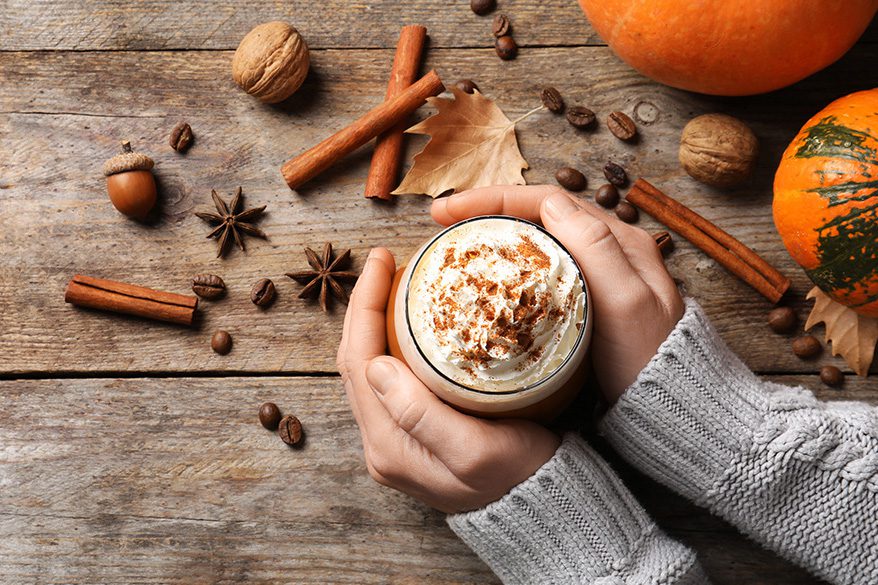 Woman's hand around a pumpkin spice latte, with spices, nuts, leaves and pumpkin